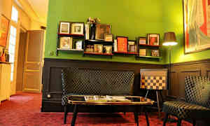 Et rom med grønne vegger og eklektisk interiør, inkludert en hylle med bøker og dekorasjoner, en sofa, og en stol med sjakkmønster. (AI-generert tekst)