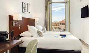 Et hotellrom med dobbeltseng, skrivebord og en åpen balkongdør som viser utsikten over omkringliggende bygninger. (AI-generert tekst)