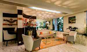 En stue med komfortable lenestoler, bord og sofa på et oransje teppe, med planter og kunst på veggene. Stor speil og vinduer. (AI-generert tekst)