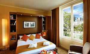 Et hotellrom med en dobbeltseng, puter og tepper i varme farger samt en stor vindusutsikt mot trær og bygninger. (AI-generert tekst)