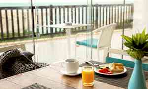 Frokostbord med kaffe, juice og grønnsaker nær et glassvindu med utsikt over en terrasse med havet i bakgrunnen. (AI-generert tekst)