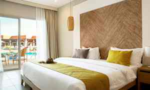 Hotellrom med dobbeltseng, gule og brune puter, trestrukturert sengegavl, og balkong med utsikt over bassenget. (AI-generert tekst)