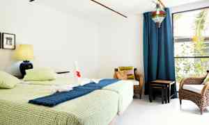 Et rom med to enkeltsenger, en blå lampe på et nattbord, to stoler av rotting og et stort vindu med blå gardiner. (AI-generert tekst)