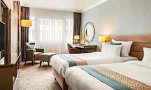 Et hotellrom med to enkeltsenger, skrivebord, stol og lenestol. (AI-generert tekst)