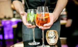 To drinker holdes frem av en person, et glass inneholder en oransje cocktail og det andre en klar cocktail med lime og mynte. (AI-generert tekst)