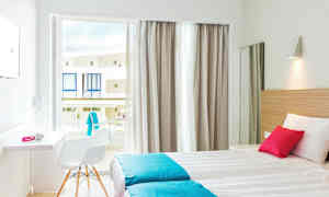 Et lyst hotellrom med to enkeltsenger, hvite og turkise sengetøy, en rosa pute. Vinduet har lyse gardiner og åpner mot balkonger. (AI-generert tekst)