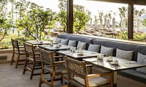 Utendørsområde med bord, stoler og benk med puter foran grøntområder og solsenger. (AI-generert tekst)