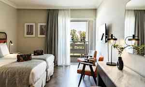 Hotellrom med to enkeltsenger, sittegruppe og skrivebord. Balkong med utsikt over trær. (AI-generert tekst)