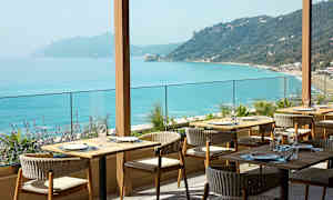 En restaurant med uteservering og utsikt over turkisblått hav og kupert kystlinje. (AI-generert tekst)