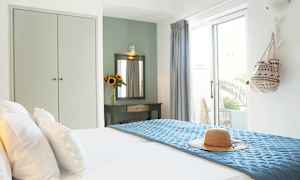 Et lyst hotellrom med en oppredd seng, en solhatt på blått sengeteppe og et dekorasjonsbord med speil og en vase med solsikker. (AI-generert tekst)