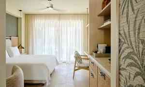 Et hotellrom med en stor seng, en lenestol og hyller med kaffemaskin, opplyst av naturlig lys gjennom store gardiner. (AI-generert tekst)