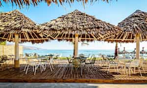 Utendørsområde med bord og stoler under stråtak nær stranden. (AI-generert tekst)