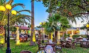 En utendørs restaurant med rottingbord og stoler, palmer og dekorerte lyslykter, omgitt av grøntområder og belysning. (AI-generert tekst)