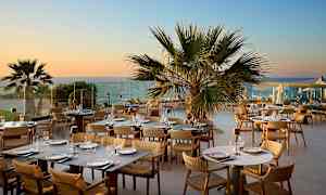 En utendørs restaurant med bord og stoler, omgitt av palmer og med utsikt over havet i solnedgangen. (AI-generert tekst)