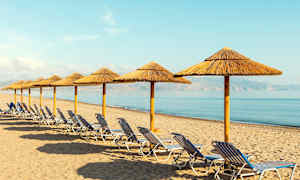 En rekke med solsenger og parasoller laget av halm på en sandstrand ved sjøen. (AI-generert tekst)