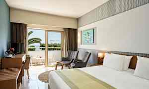 Et hotellrom med en dobbeltseng, to stoler, et bord og en TV-møbel. En balkong med utsikt mot palmer er synlig gjennom en glassdør. (AI-generert tekst)