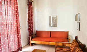 Stuen har røde gardiner, en tresofa med rødt rutete puter, og en stol med tekstiltrekk ved siden av. (AI-generert tekst)