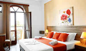 Et hotellrom med en seng kledd i hvite og oransje laken, et tre-skrivebord og en åpning mot en balkong. (AI-generert tekst)