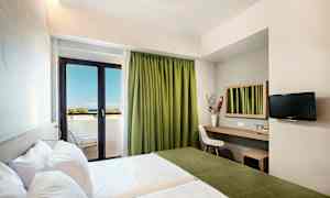 Et hotellrom med to senger, en arbeidsflate med speil, en stol og en TV på veggen. Rommet har en balkong med grønne gardiner og utsikt mot havet. (AI-generert tekst)