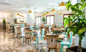 En restaurant med bord og stoler, samt store vinduer og grønne planter. (AI-generert tekst)