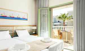 Et lyst hotellrom med to enkeltsenger, hvitt sengetøy og beige sengetepper. En kunst med båt motiver henger på veggen over sengen. Rommet har en åpen balkongdør med utsikt over en palme og et bord med to stoler. (AI-generert tekst)