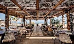 Utendørs servering med trebord og stoler under tak med utsikt over havet. (AI-generert tekst)