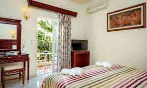 Et hotellrom med seng, TV, sminkbord og balkong med utsikt mot en palme. (AI-generert tekst)