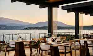 En restaurant med trebord og stoler, plassert ved sjøen med utsikt over fjell i bakgrunnen under solnedgangen. (AI-generert tekst)