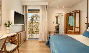 Hotellrom med dobbeltseng, skrivebord og TV. Glassdører fører ut til en balkong. Baderommet er synlig gjennom en åpen dør. (AI-generert tekst)