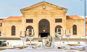 Et dekket bord utendørs med vinglass, bestikk og servietter. I bakgrunnen sees en historisk steinbygning. (AI-generert tekst)