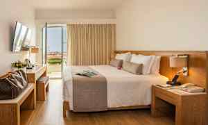 Et hotellrom med en stor dobbeltseng, skrivebord og åpne dører mot en balkong med utsikt. (AI-generert tekst)