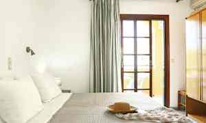 Et soverom med en seng, puter og et teppe. Et vindu med gardiner og et nattbord er synlig. (AI-generert tekst)