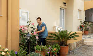 Person tar vare på blomster ved et hus med beige vegger. (AI-generert tekst)