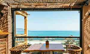 En uteplass med bord og stoler, utsikt over havet og stranden gjennom en ramme. (AI-generert tekst)