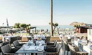 Utendørs servering med bord og stoler, havutsikt og strand i bakgrunnen. (AI-generert tekst)