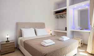 Et hotellrom med en dobbeltseng utstyrt med beige sengeteppe og to sett med håndklær, samt sidebord og garderobe. (AI-generert tekst)