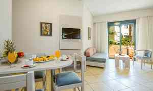 Et lyst hotellrom med spisebord og bakverk på en tallerken, sofagruppe og en balkong i bakgrunnen. (AI-generert tekst)