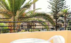 Utsikt fra balkong med et bord og plaststol, palmetrær og bygninger i bakgrunnen. (AI-generert tekst)