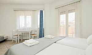 Et lyst rom med to enkeltsenger, et bord med to stoler og vinduer dekket av hvite gardiner. (AI-generert tekst)