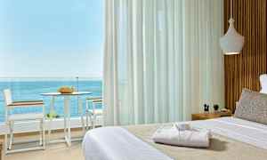 Utsikt fra et rom med en oppredd seng mot en balkong med stoler, bord og havutsikt. (AI-generert tekst)