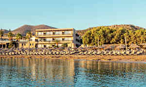 En ferieanlegg ved stranden med solsenger og parasoller i forgrunnen og et hotell bak. (AI-generert tekst)