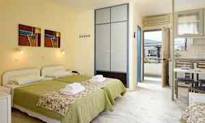 To senger med grøn sengetøy og blomstrete sengeoverkast i et rom med skap, kjøkkenbord og dør til balkong. (AI-generert tekst)