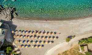 Luftbilde av en strand med oppstilte solsenger og parasoller ved kanten av turkist vann. (AI-generert tekst)