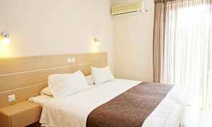 Et hotellrom med en stor seng, hvitt sengetøy og brunt overkast. En klimaanlegg er montert på veggen ved siden av vinduet med lyse gardiner. (AI-generert tekst)