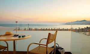 Utsikt fra en terrasse mot havet med solnedgang og strandparasoller i bakgrunnen. (AI-generert tekst)