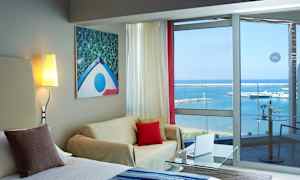 Et hotellrom med sittegruppe, lysmaleri på veggen og glassvinduer med utsikt over havet og en småbåthavn. (AI-generert tekst)