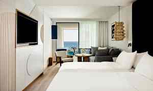 Et hotellrom med to senger, sittemøbler og en balkong med utsikt over havet. (AI-generert tekst)