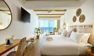 Et hotellrom med hvite vegger, en oppredd seng, sittemøbler og en balkong med utsikt over havet. (AI-generert tekst)