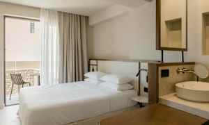 Interiør av et hotellrom med en dobbeltseng, et vindu med gardiner, en balkong med stol og en vask. (AI-generert tekst)