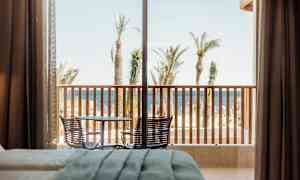 Hotellrom med utsikt over en terrasse med bord og stoler, etterfulgt av palmer og havet i bakgrunnen. (AI-generert tekst)